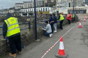 A photo of Ilfracombe town, with a collection of people cleaning a fence together. One of them wears a hi-vis jacket with 'Ilfracombe Town Council' on the back